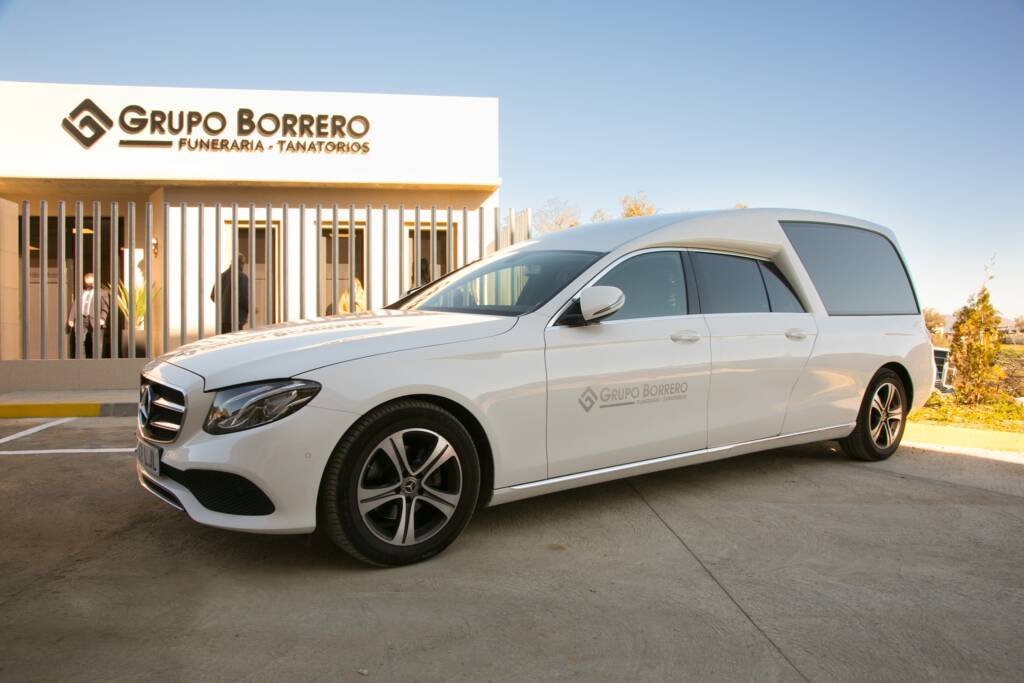 Un coche fúnebre blanco está aparcado delante de un edificio con un letrero que dice Grupo Borrero Funeraria - Tanatorios. El edificio tiene barrotes verticales en las ventanas y se ve un cielo despejado.
