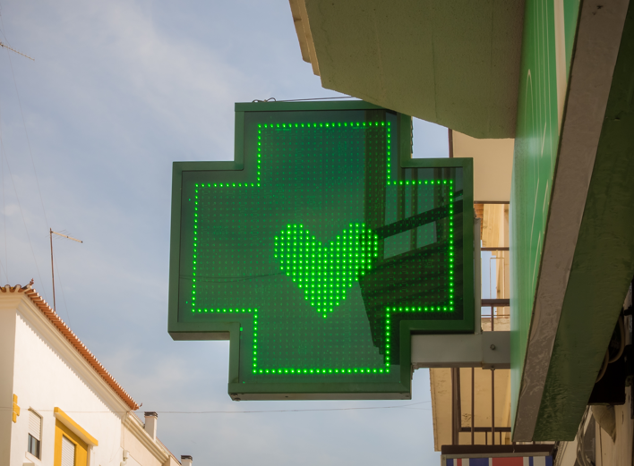Una señal LED verde en forma de cruz con el símbolo de un corazón en el centro está montada en el exterior de un edificio, indicando la ubicación de una farmacia. El cielo y partes de edificios vecinos son visibles.