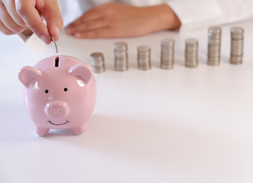 Una persona deposita una moneda en una hucha rosa, con varias pilas de monedas dispuestas al fondo sobre una superficie blanca.