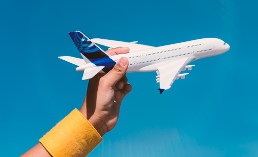 Una mano con una muñequera amarilla sostiene un avión de juguete blanco con cola azul sobre un cielo azul despejado.